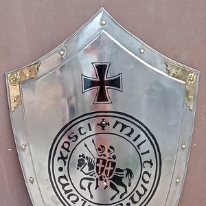 May include: A polished silver-tone metal heater shield featuring a black iron cross at the top, a central circular emblem depicting two knights on a horse with the text "xpsti militum sigillum", and a black Celtic knot design at the bottom.