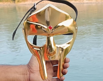 May include: A gold-colored metal mask with a gladiator design. The mask has a red jewel on the forehead and a black leather strap.