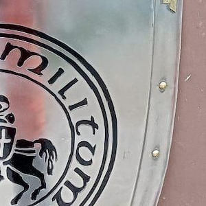 May include: A polished silver-toned metal shield featuring a circular black emblem with a horse and shield graphic, surrounded by the text "militum" and "si". The shield has a decorative brass-colored corner piece and small rivets along the edge.