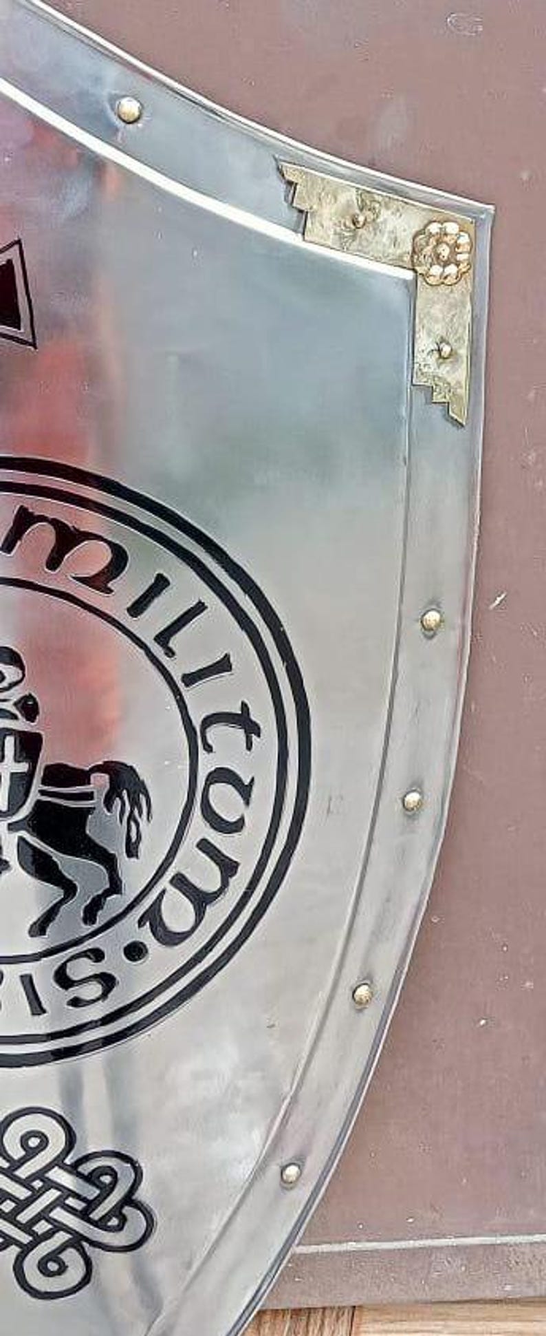 May include: A polished silver-toned metal shield featuring a circular black emblem with a horse and shield graphic, surrounded by the text "militum" and "si". The shield has a decorative brass-colored corner piece and small rivets along the edge.