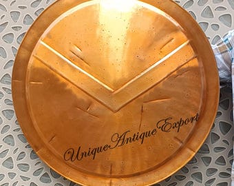 May include: A round, copper-colored shield with a V-shaped design in the center. The shield has a slightly textured surface and the words "Unique Antique Export" are written in cursive script.