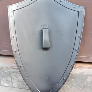 May include: A pointed heater-style shield with a smooth matte black finish. The shield features a raised border along the perimeter secured with evenly spaced metal rivets. A vertical rectangular handle is mounted in the center of the shield.