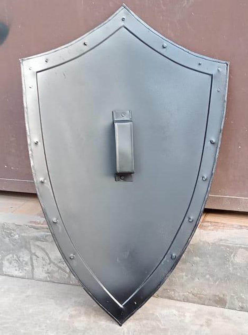 May include: A pointed heater-style shield with a smooth matte black finish. The shield features a raised border along the perimeter secured with evenly spaced metal rivets. A vertical rectangular handle is mounted in the center of the shield.