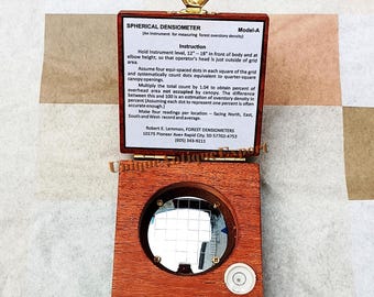 May include: A Spherical Densiometer Model-A, a wooden instrument for measuring forest overstory density. The device has a circular mirror with a grid, a level, and a hinged lid with instructions. The text on the lid includes the manufacturer's address.