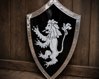 May include: A heater-style shield with a glossy black finish and a silver metallic border secured by rivets. The centre features a white graphic illustration of a rampant lion with its tongue extended, depicted in a classic heraldic style.