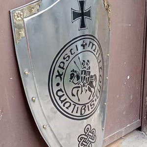 May include: A polished silver-tone metal heater shield featuring a black iron cross at the top, a central circular emblem depicting two figures on horseback, and a black Celtic knot design at the bottom, accented with gold-tone corner plates.