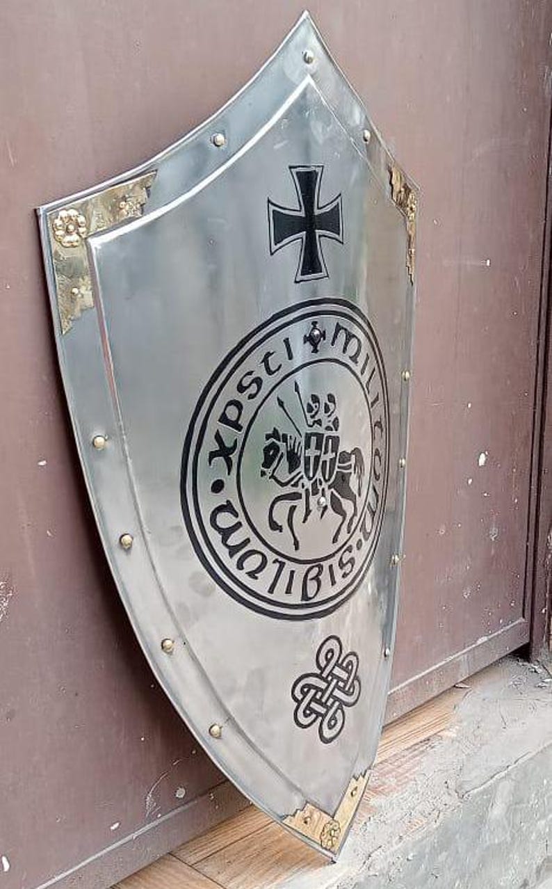 May include: A polished silver-tone metal heater shield featuring a black iron cross at the top, a central circular emblem depicting two figures on horseback, and a black Celtic knot design at the bottom, accented with gold-tone corner plates.