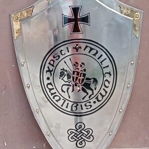 May include: A polished silver-tone metal shield featuring a central black graphic of two knights on a horse, surrounded by the text Sigillum Militum Xpsti. Above is a black cross, and below is a decorative knot design with gold-tone corner accents.