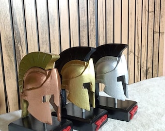May include: Three Spartan helmet replicas mounted on black rectangular bases. The helmets are finished in metallic copper, gold, and silver, each featuring a black crest. The front of each black base displays the red text 300 in a stylized font.