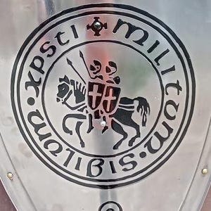 May include: A polished silver-toned metal shield featuring a black Iron Cross at the top. The center displays a circular emblem with the Latin text 'Sigillum Militum Xpsti' surrounding an engraving of two knights on a single horse. A knot design sits below.