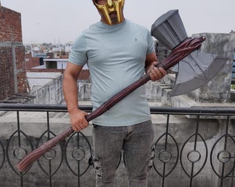 May include: A gold-colored metal mask with a distinct facial design, paired with a large, long-handled battle axe featuring a silver-toned head and a dark brown, textured handle. The axe head has a hammer-like top and a sharp, wide blade.