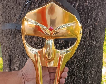 May include: A gold-colored mask with a reflective surface, featuring a prominent nose and mouth area. A red gem is set above the nose. The mask has a black strap attached. The mask is held in a hand.