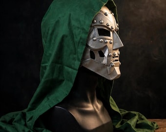 May include: A silver-colored metallic mask with a riveted design and rectangular eye openings, worn on a black mannequin head. The mask is partially covered by a dark green fabric hood that drapes down over the shoulders of the mannequin.