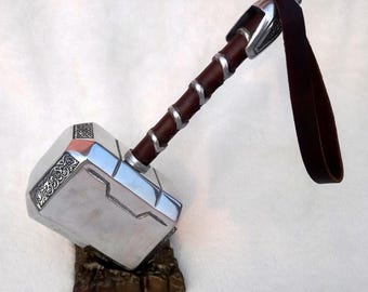 May include: A silver and brown replica of Thor's hammer, Mjolnir, with intricate detailing. The hammer features a brown leather handle wrapped with silver accents and a matching leather strap. It rests on a brown, textured base.