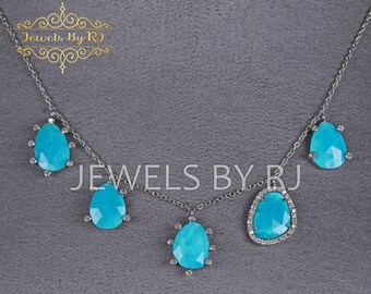 May include: A silver chain necklace with five turquoise teardrop-shaped stones. The center stone is surrounded by a halo of smaller stones. The necklace is on a gray background. "JEWELS BY RJ" is written in the top left corner of the image.