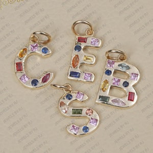May include: Four gold-toned metal letter charms shaped as C, E, B, and S. Each letter is inlaid with various multi-coloured gemstones in different shapes, including round, square, rectangular, and marquise cuts. Each charm features a small circular jump ring.