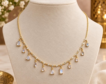 May include: A gold-toned necklace featuring a delicate chain adorned with small, clear round stones and multiple dangling charms. The charms consist of various geometric shapes, including rectangles, triangles, and teardrops, each set with a translucent, iridescent stone.