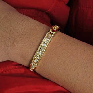 May include: A gold bangle bracelet with a row of clear cubic zirconia stones set in a pave setting. The bracelet is open and has a hinged clasp.