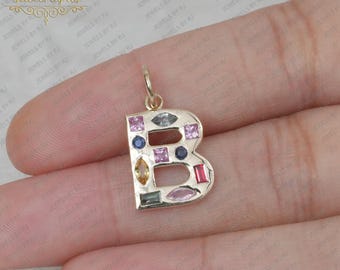 以下が含まれることがあります： 指に置かれた、Bの字の形をした金のチャームペンダント。カラフルな宝石が文字に埋め込まれている。