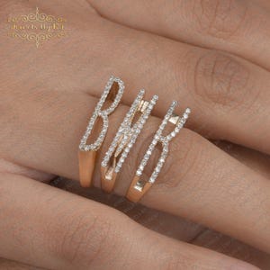 Op de afbeelding: Een roségouden ring met de letters "BHR" ingelegd met kleine, sprankelende diamanten. De ring heeft een uniek, open ontwerp met de letters verticaal gerangschikt. De ring zit om de vinger van een persoon.
