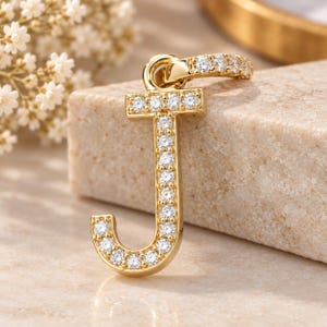 May include: A gold-toned letter J pendant featuring a series of clear, round-cut stones embedded along the front surface. The pendant includes a small, circular bail at the top, which is also adorned with a row of matching clear, round-cut stones.