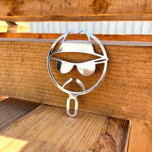 May include: A shiny, silver metal cutout of a fedora-wearing figure with sunglasses and a mustache. The cutout is circular with a handle at the bottom. The object is resting on a wooden surface.
