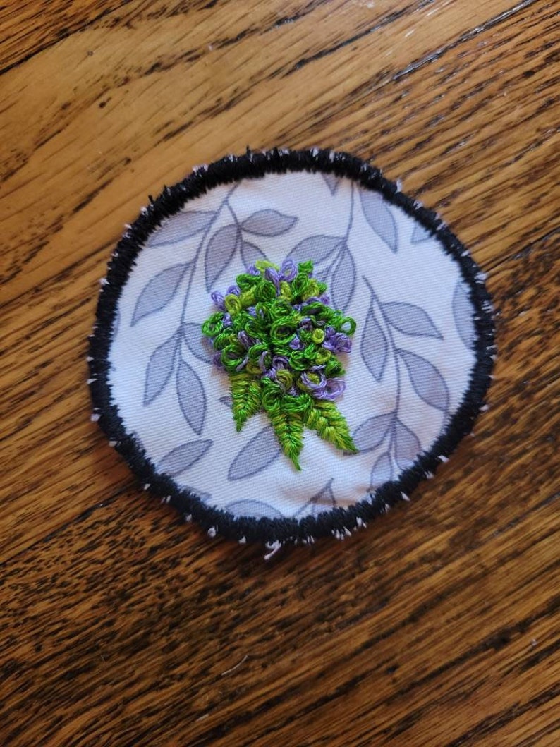 May include: A round embroidered patch featuring a purple and green flower design on a white background with a grey leaf pattern. The patch has a black border.