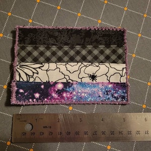 May include: A purple, black, white and blue fabric patch with a galaxy print, floral print and chequered pattern. The patch is sewn with a zig-zag stitch around the edges.