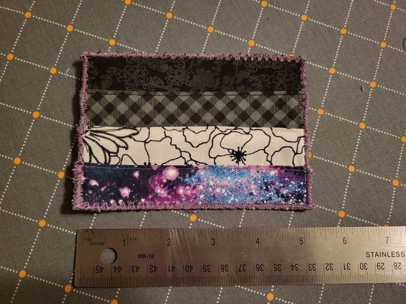 May include: A purple, black, white and blue fabric patch with a galaxy print, floral print and chequered pattern. The patch is sewn with a zig-zag stitch around the edges.