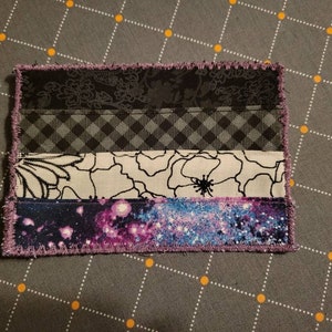 May include: A handmade fabric patch with a purple, black, white and grey design. The patch is made of different fabrics sewn together to create a layered effect. The patch has a fringed edge.