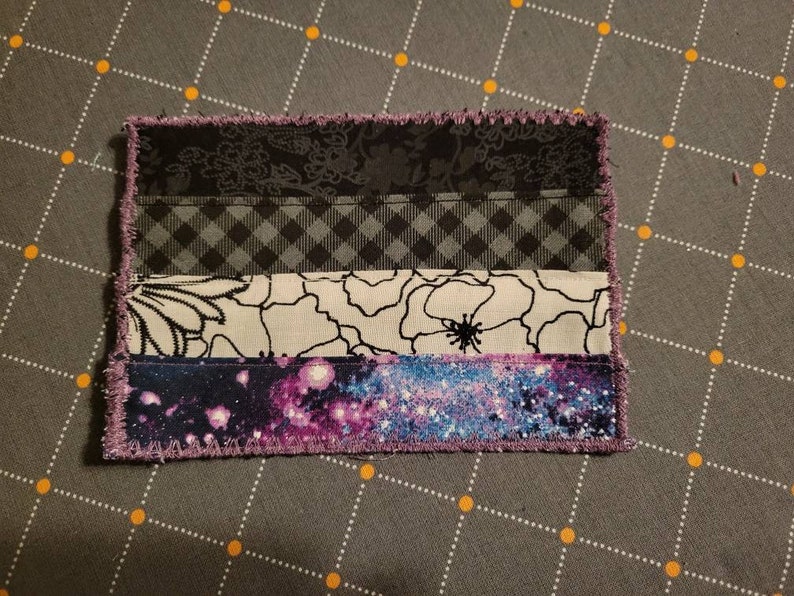 May include: A handmade fabric patch with a purple, black, white and grey design. The patch is made of different fabrics sewn together to create a layered effect. The patch has a fringed edge.