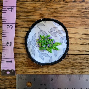 May include: A round, white fabric patch with a black border. The patch features a floral design with green leaves and purple flowers embroidered on a white background with a grey leaf pattern.