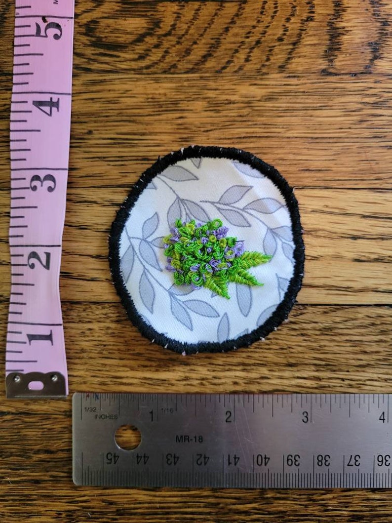 May include: A round, white fabric patch with a black border. The patch features a floral design with green leaves and purple flowers embroidered on a white background with a grey leaf pattern.