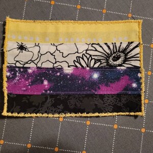 May include: A handmade non-binary pride flag patch with yellow, white, purple, and black stripes. The patch is made of fabric with a galaxy print and floral designs.
