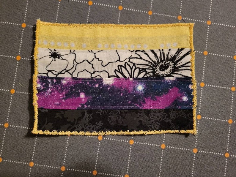 May include: A handmade non-binary pride flag patch with yellow, white, purple, and black stripes. The patch is made of fabric with a galaxy print and floral designs.