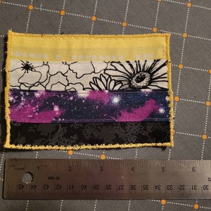May include: A small, handmade flag with five horizontal stripes. The top stripe is yellow, the second is white with black floral designs, the third is a purple and blue galaxy print, the fourth is black, and the bottom stripe is yellow. The flag is sewn with a yellow zig-zag stitch around the edges.