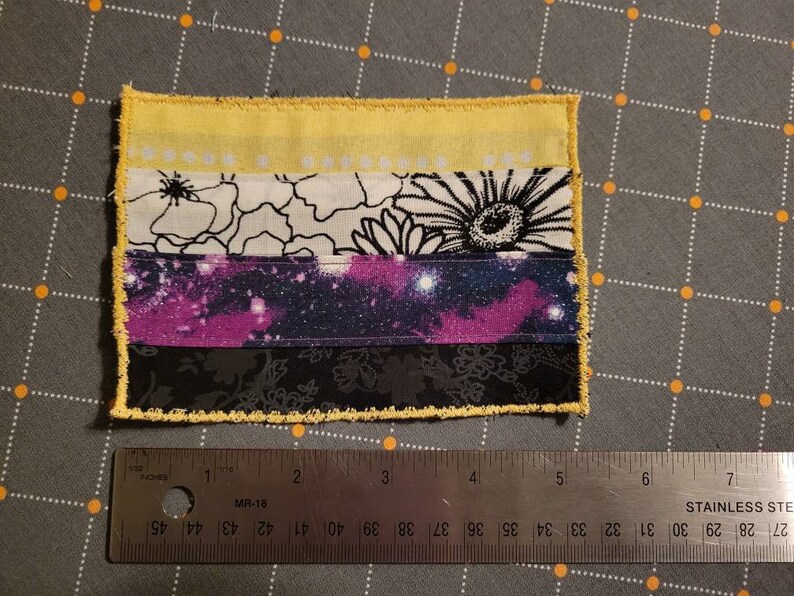 May include: A small, handmade flag with five horizontal stripes. The top stripe is yellow, the second is white with black floral designs, the third is a purple and blue galaxy print, the fourth is black, and the bottom stripe is yellow. The flag is sewn with a yellow zig-zag stitch around the edges.