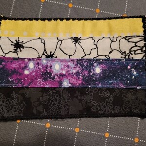 May include: A handmade fabric patch with a yellow, white, black, purple, and black color scheme. The patch is made of different fabrics with various patterns, including floral and galaxy prints. The patch is edged with black stitching.