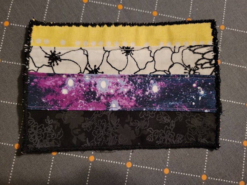 K&ouml;nnte beinhalten: Ein handgefertigter Stoffpatch mit einem Farbschema aus Gelb, Wei&szlig;, Schwarz, Lila und Schwarz. Der Patch besteht aus verschiedenen Stoffen mit verschiedenen Mustern, darunter Blumen- und Galaxie-Drucke. Der Patch ist mit schwarzen N&auml;hten eingefasst.