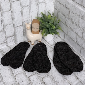 May include: Three pairs of black wool insoles on a white brick background. The insoles are made of a soft, fuzzy material and are designed to provide comfort and support.