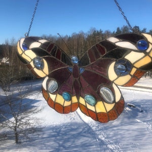 May include: A stained glass butterfly with a purple, black, white, and yellow design. The butterfly has blue and clear glass accents. The butterfly is hanging from a chain.