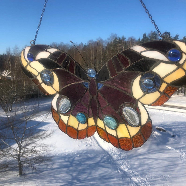 May include: A stained glass butterfly with a purple, black, white, and yellow design. The butterfly has blue and clear glass accents. The butterfly is hanging from a chain.