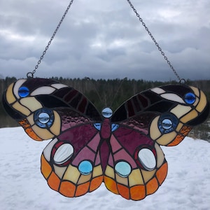 May include: A stained glass butterfly window hanging with a black metal chain. The butterfly has a purple, orange, and yellow body with black and white wings. The wings have blue and white accents.