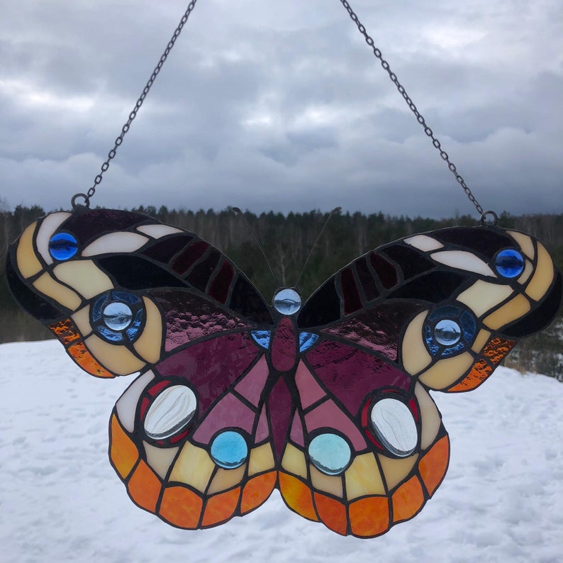 May include: A stained glass butterfly window hanging with a black metal chain. The butterfly has a purple, orange, and yellow body with black and white wings. The wings have blue and white accents.