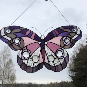 May include: A stained glass butterfly with purple, pink, and brown accents. The butterfly has clear glass accents and is hanging from a silver chain.