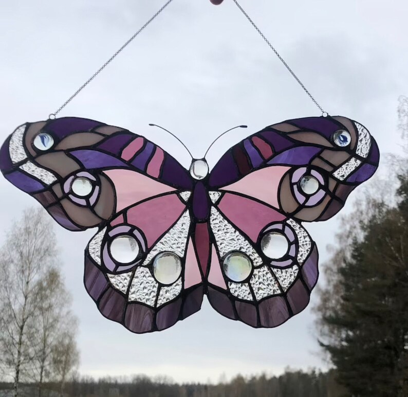 May include: A stained glass butterfly with purple, pink, and brown accents. The butterfly has clear glass accents and is hanging from a silver chain.