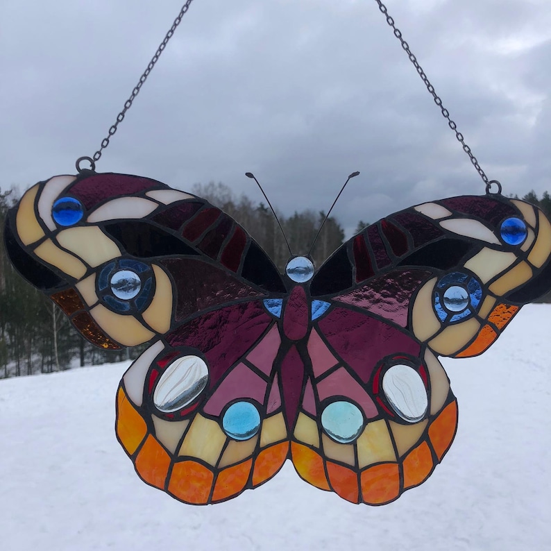 May include: A stained glass butterfly suncatcher with a purple, orange, and brown design. The butterfly has blue and white accents and is hanging from a chain.