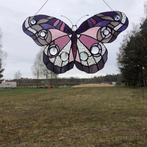 May include: A stained glass butterfly with purple, pink, and white accents. The butterfly is hanging from a string and is in front of a field.