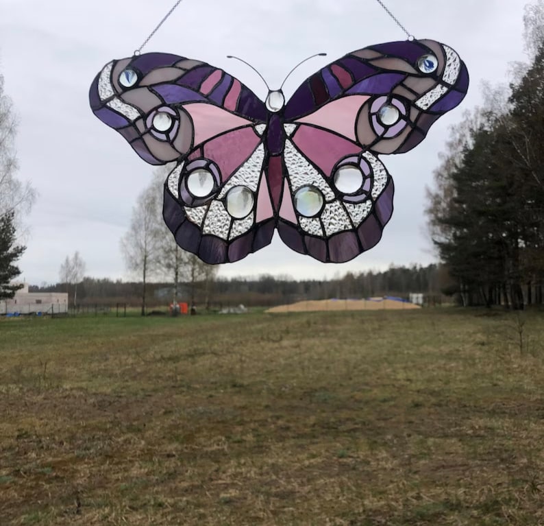 May include: A stained glass butterfly with purple, pink, and white accents. The butterfly is hanging from a string and is in front of a field.
