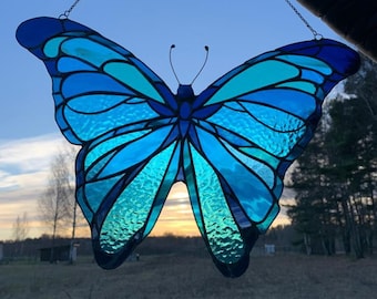 May include: A stained glass butterfly with blue and teal glass, hanging from a silver chain against a blue sky background.